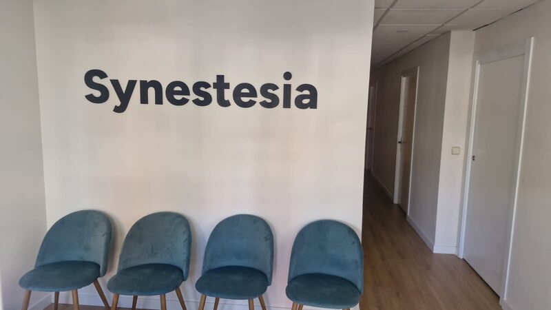 El rótulo Synestesia en letras oscuras sobre una pared blanca, encima de cuatro sillas azules en la sala de espera de la clínica.